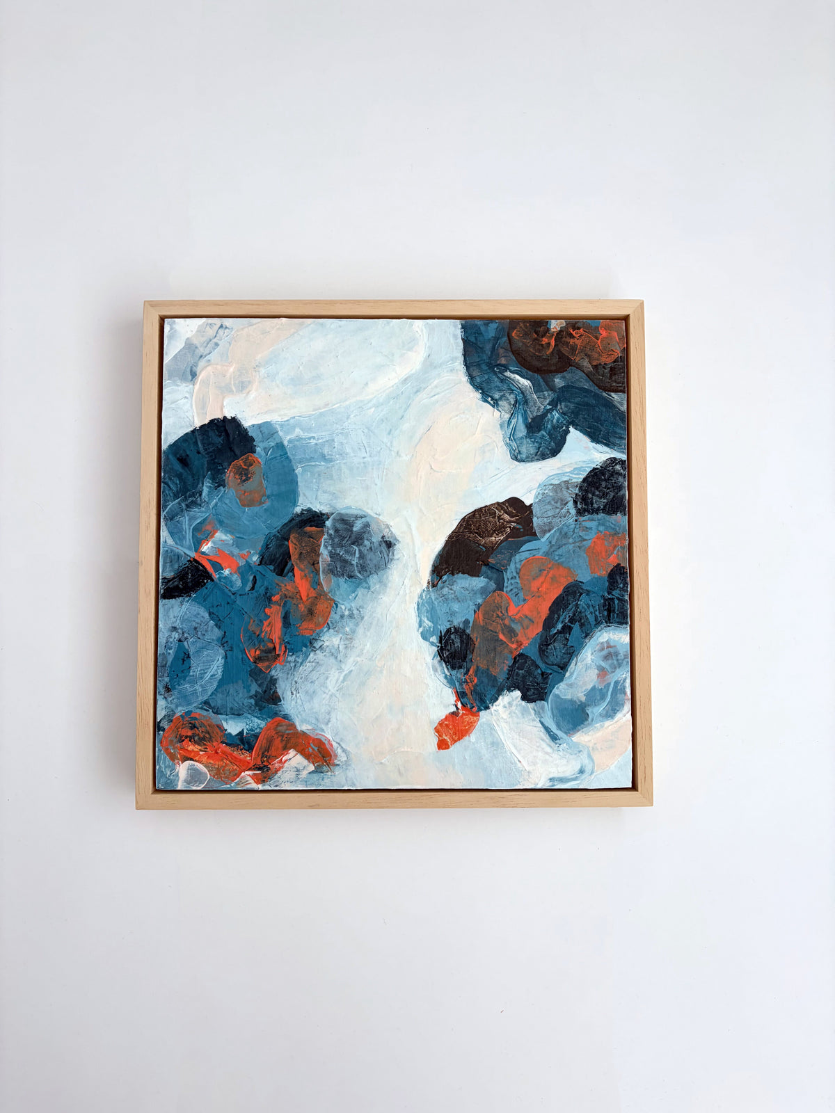An abstract framed painting featuring textured brushstrokes of blue, white, brown, and orange, displayed on a clean white wall.