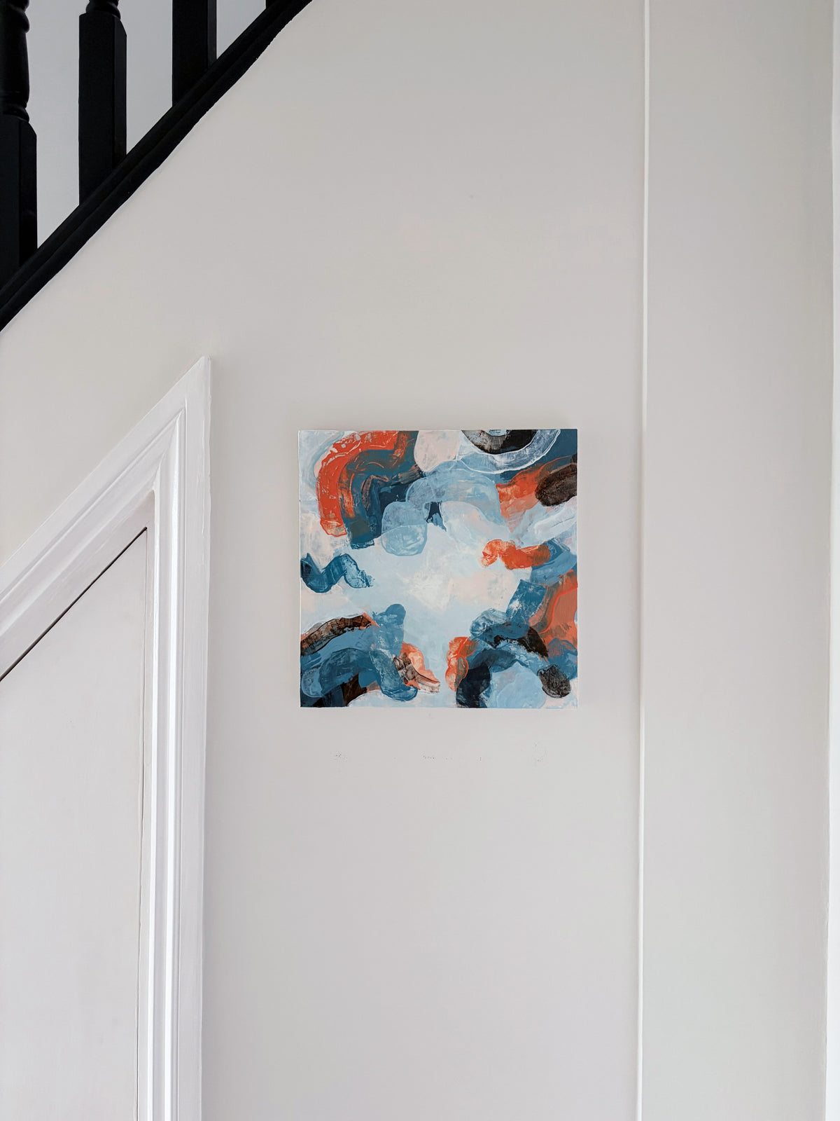 An abstract panel painting with blue, orange, and white brushstrokes hanging on a white wall next to a staircase.