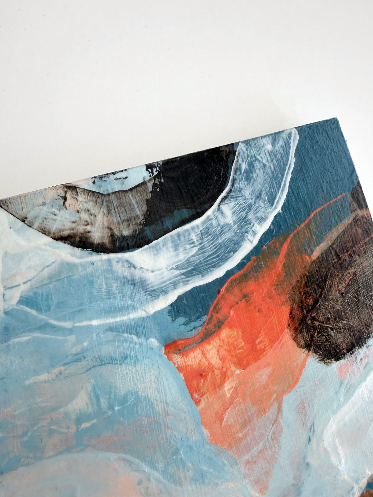 An abstract panel painting featuring textured brushstrokes of light blue, white, dark blue, and a vibrant orange-red accent, shown at an angle against a plain white background.